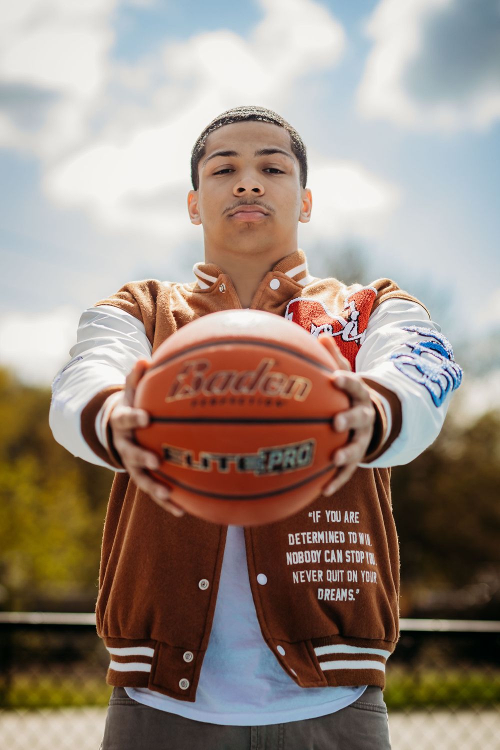 A basketball player poses confidently with a ball while wearing a varsity jacket against cloudy sky.
