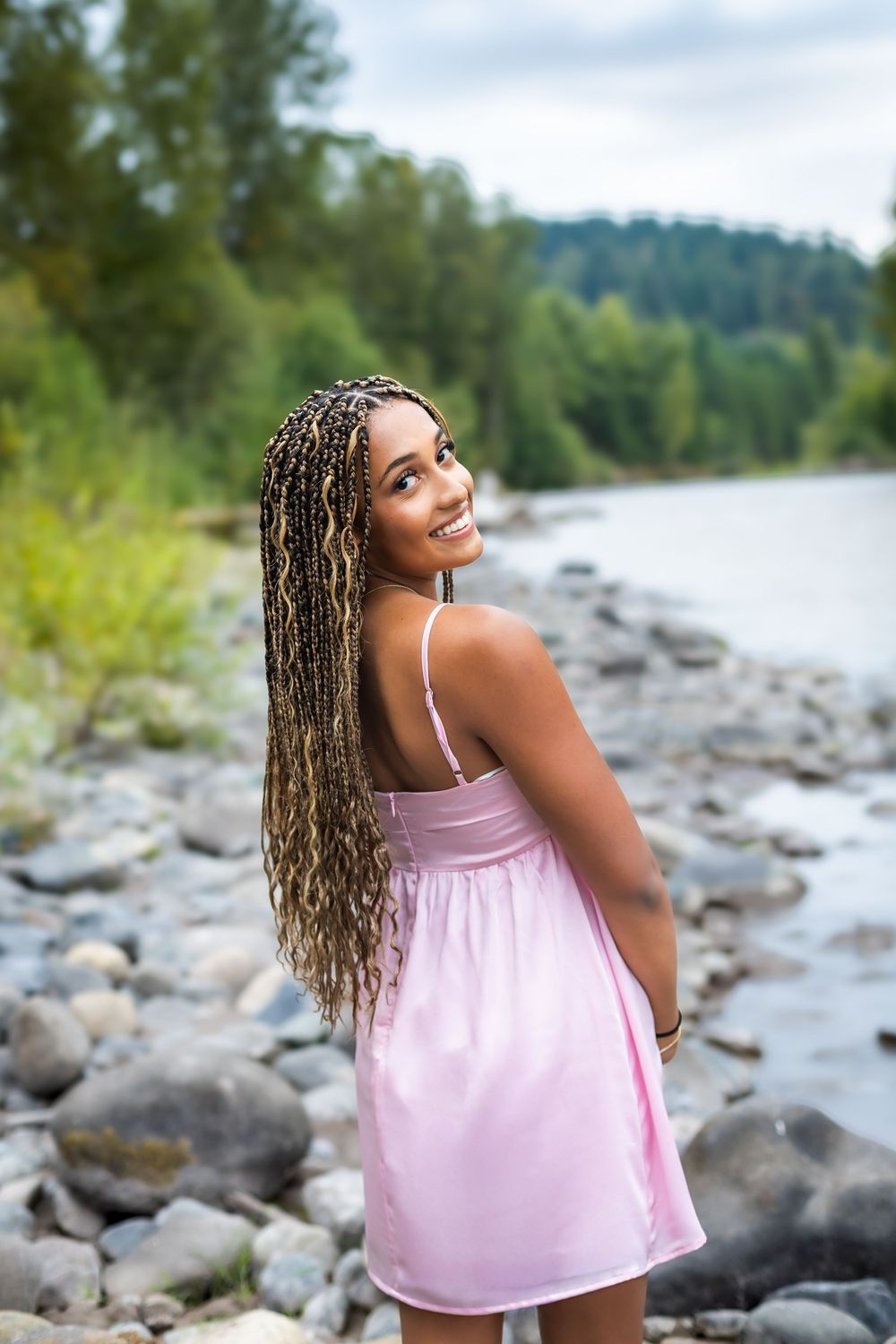 A person in a pink tank top poses on a rocky riverbank surrounded by trees and water on a cloudy day.