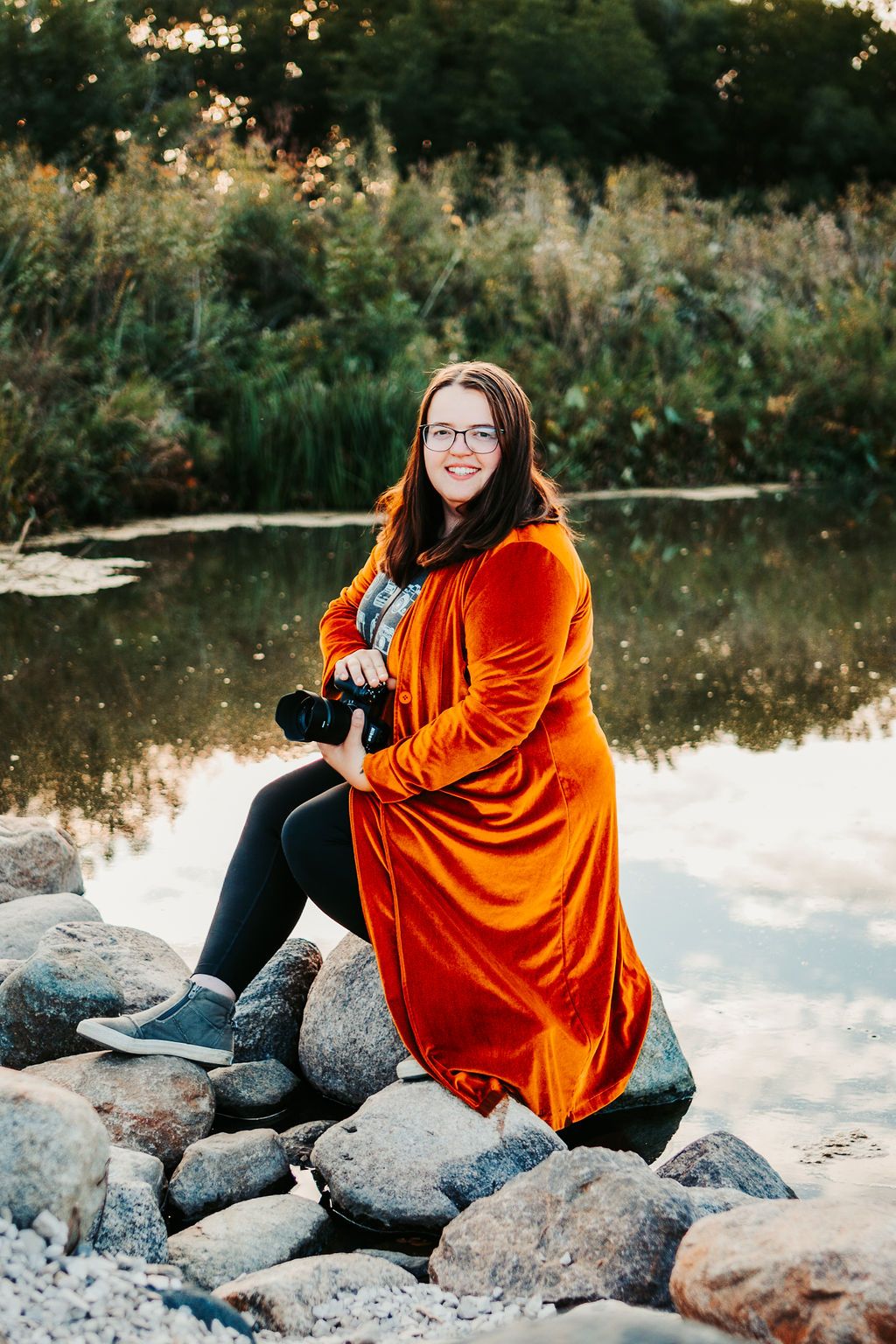 Photographer in bright orange dress sits on rocks by reflective pond at sunset.