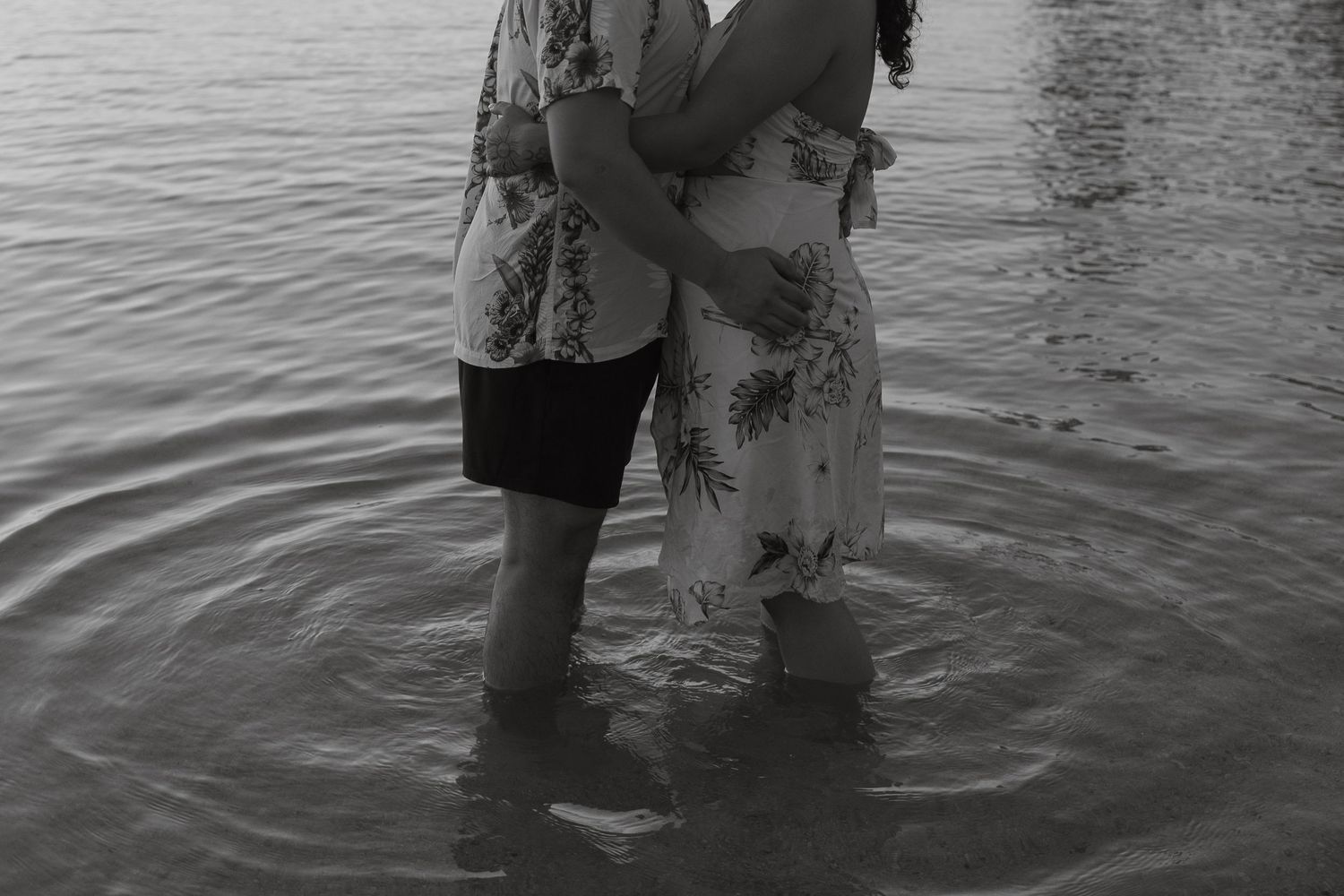Black and white photo of a couple embracing while standing in shallow water, wearing summer outfits.