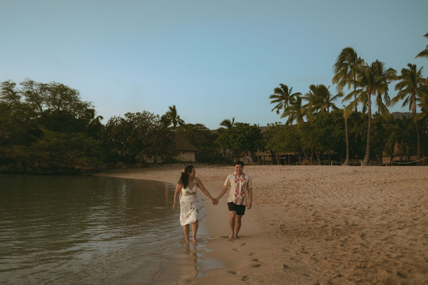 Couple holding hands walking on a tropical sandy beach at dusk with palm trees in the background.