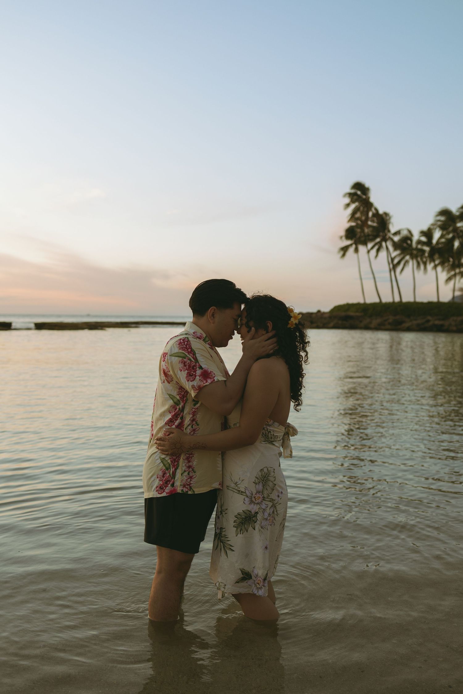 Couple in tropical attire share a romantic moment standing in shallow ocean water at sunset with palm trees.