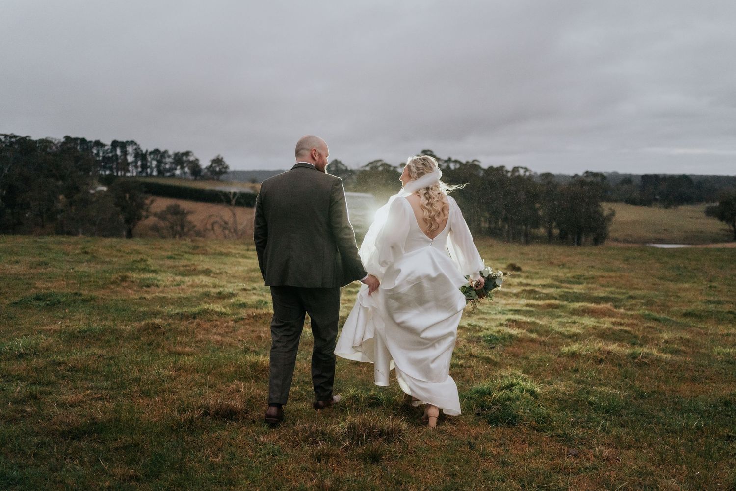 Bride and groom walking hand in hand across lush green fields on their wedding day in a rustic countryside setting.