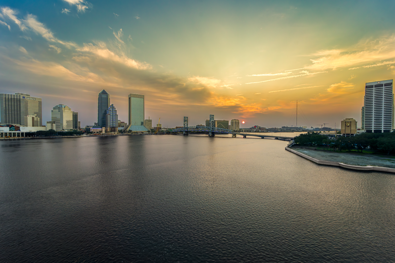 Jacksonville FL skyline at sunrise with orange sky reflecting on calm waters of St Johns River. Photo taken June 2, 2025