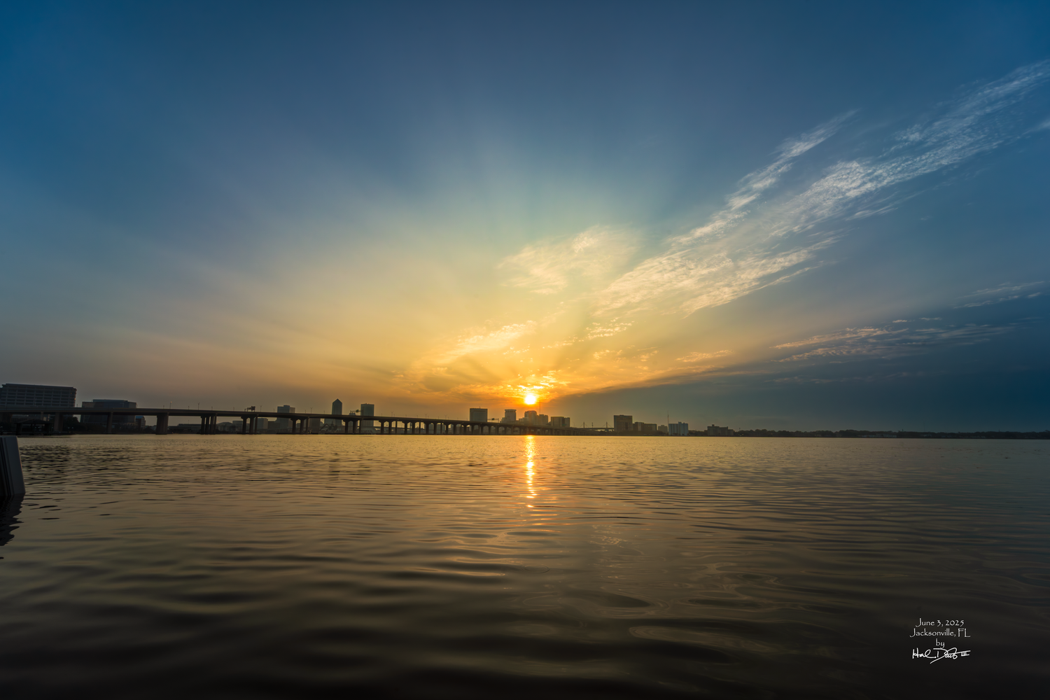 Stunning Jacksonville FL sunrise over calm water with silhouetted cityscape against dramatic golden sky and clouds. Photo taken June 3, 2025
