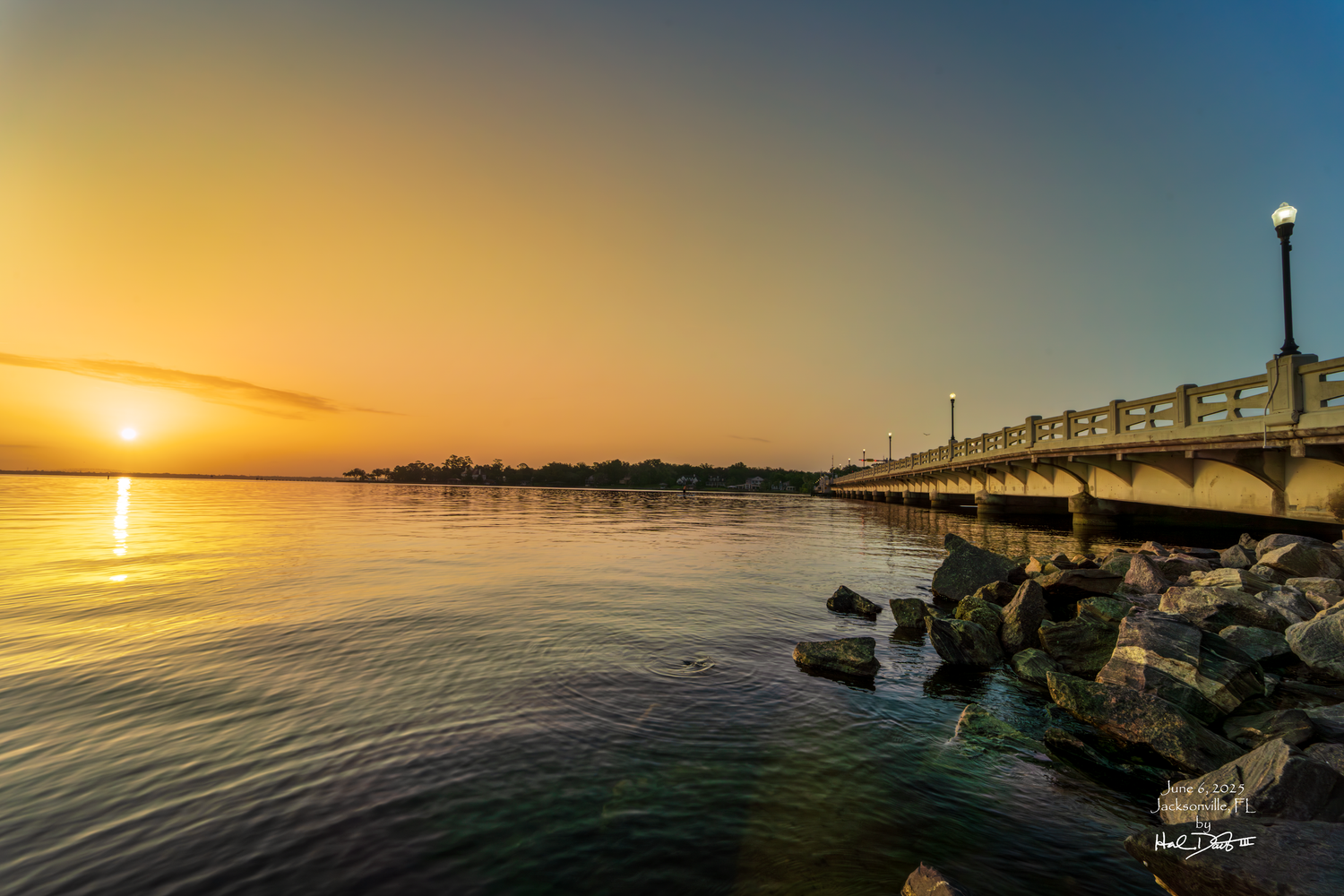Golden sunrise over calm waters with a bridge extending out over rocky shoreline at Jacksonville FL. Photo taken June 6, 2025