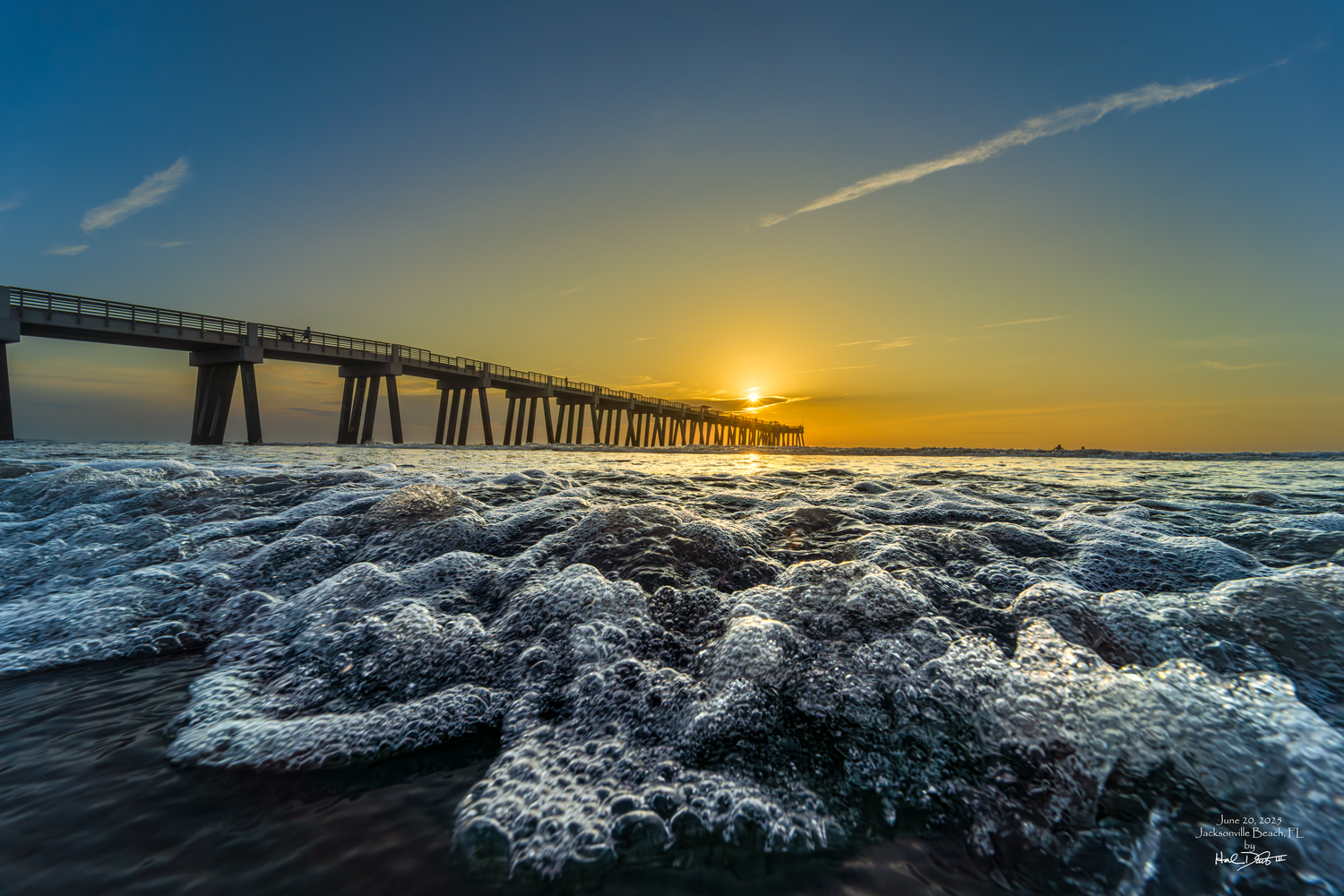 Dramatic ocean waves crash against rocks at sunset with a wooden pier silhouetted against an orange and blue sky. Photo taken June 20, 2025