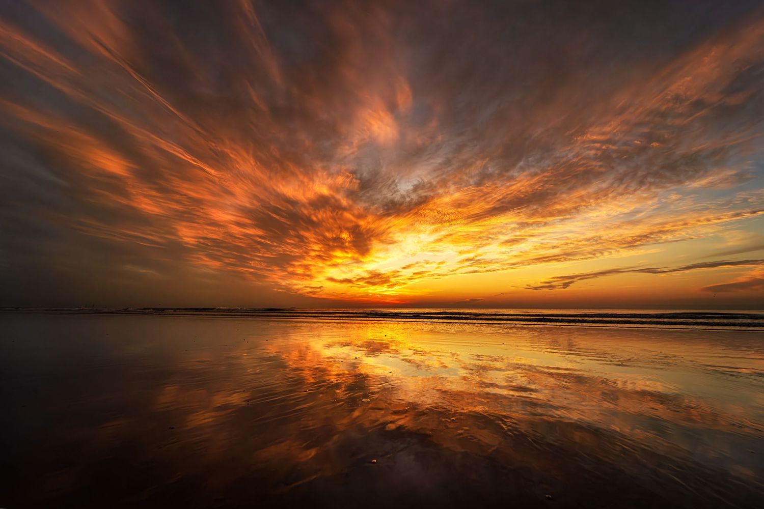 Dramatic orange and yellow sunset reflects perfectly on calm beach waters with dark storm clouds streaking across the sky.