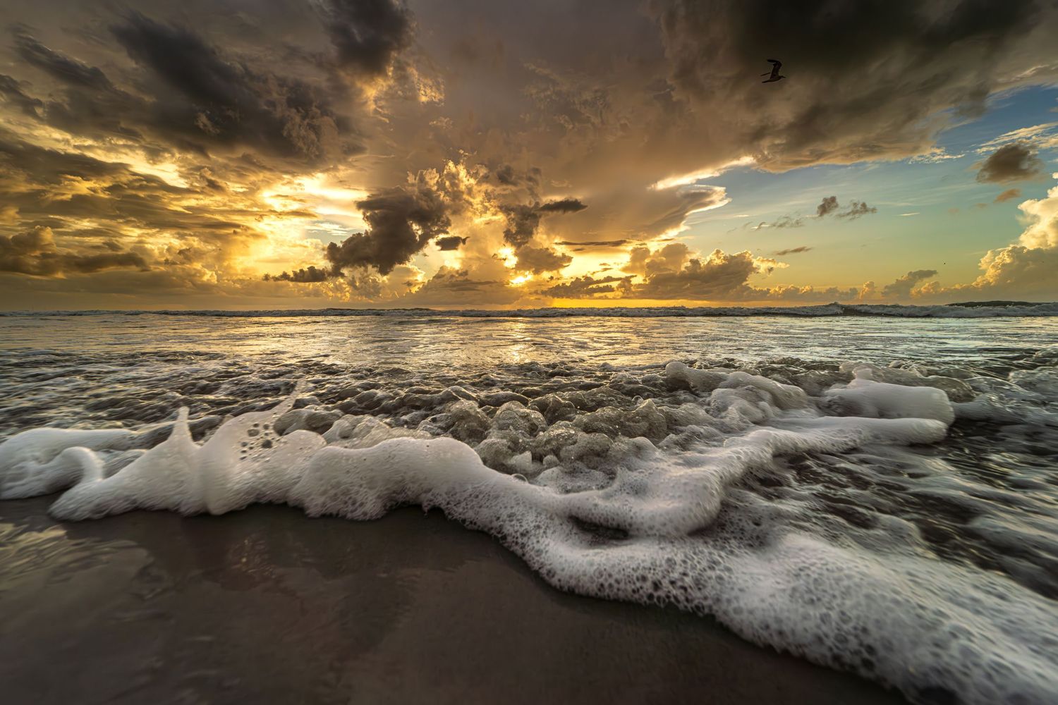 Dramatic sunrise over ocean waves with white sea foam washing ashore beneath stormy clouds and golden light.
