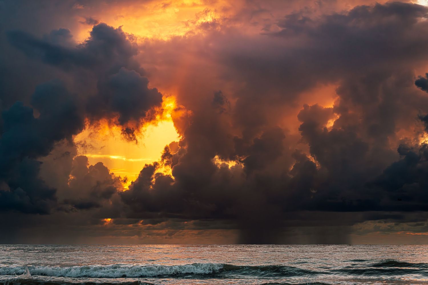 Dramatic orange sunrise breaks through dark storm clouds over turbulent ocean waves.