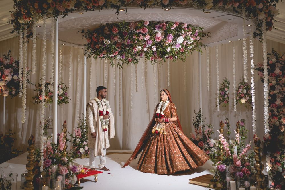 Wedding couple standing under an ornate floral mandap with hanging decorations and roses. Asian Wedding Photography & Videography