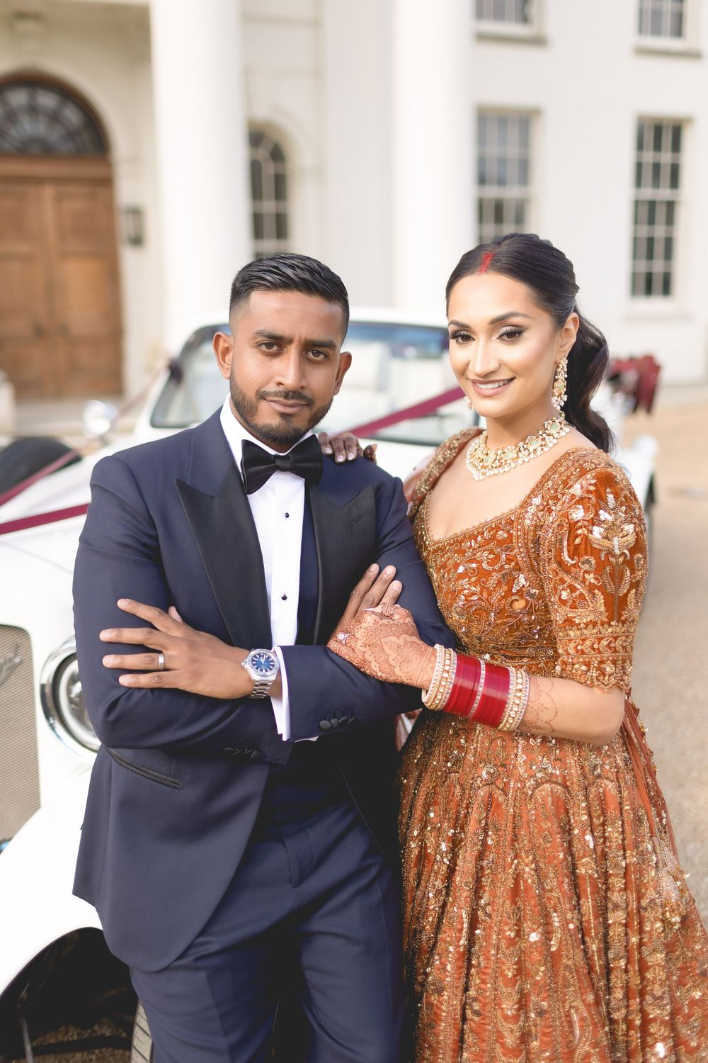 A couple in formal attire poses against a classic white car, displaying traditional wedding fashion. Asian Wedding Photography & Videography