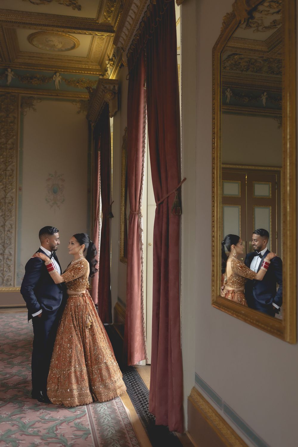 A romantic couple's reflection appears in an ornate gold mirror as they pose in a luxurious venue. Asian Wedding Photography & Videography