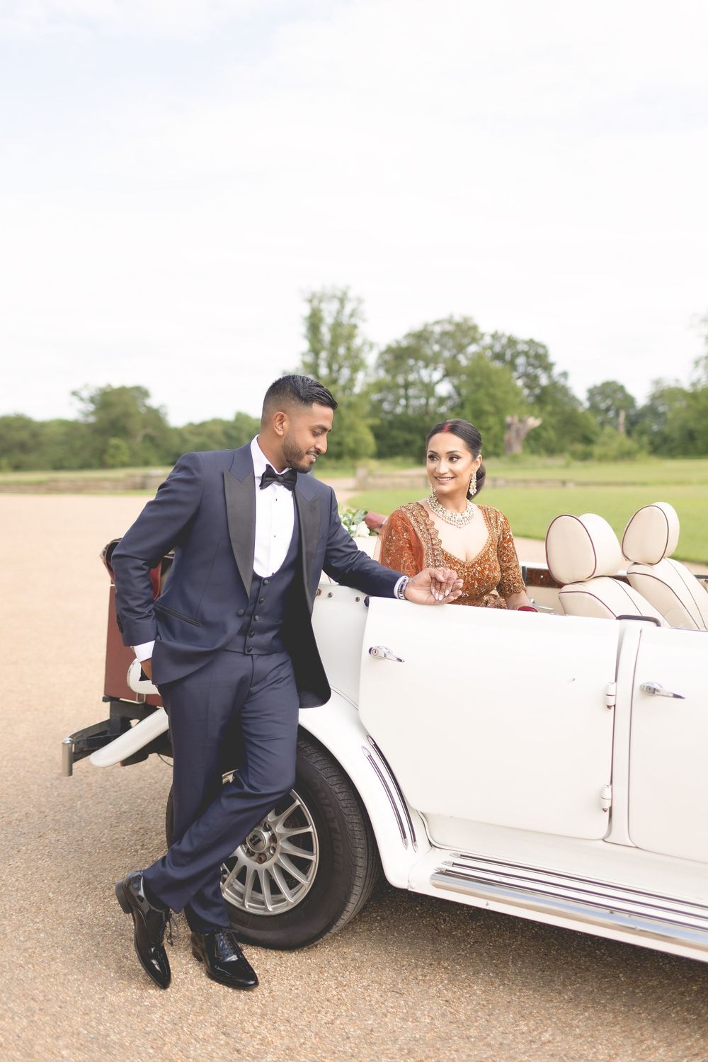 A couple poses with a classic white vintage car on a country road, showcasing elegant formal attire. Asian Wedding Photography & Videography