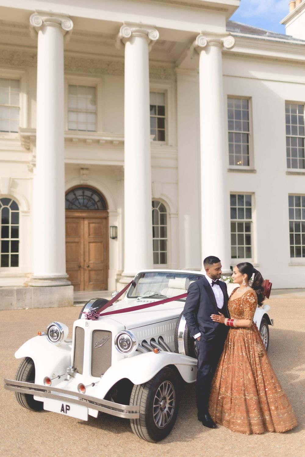 Elegantly dressed couple poses beside a vintage white car in front of a stately colonial mansion. Asian Wedding Photography & Videography