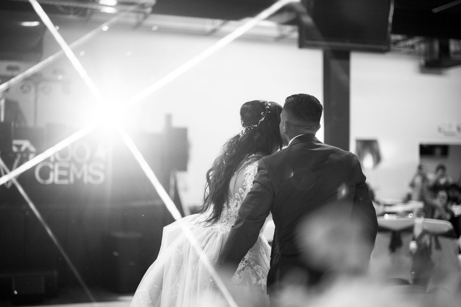A romantic black and white shot of a couple dancing at their wedding reception with dramatic backlighting creating lens flare.