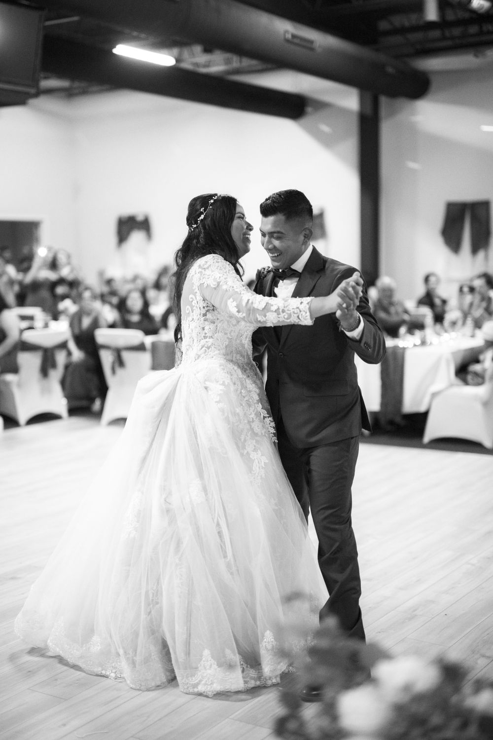 A couple shares their first dance at a wedding reception in a white decorated ballroom, captured in black and white.