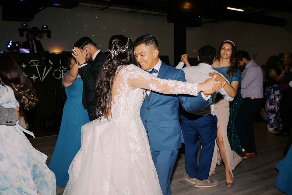 People dancing at a celebration with someone in a pink formal dress and blue suit in the foreground on a dark dance floor.