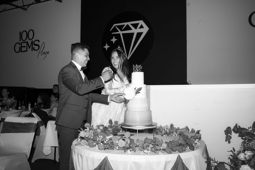 A couple in formal attire cuts their wedding cake together under a large diamond decoration at a reception venue.