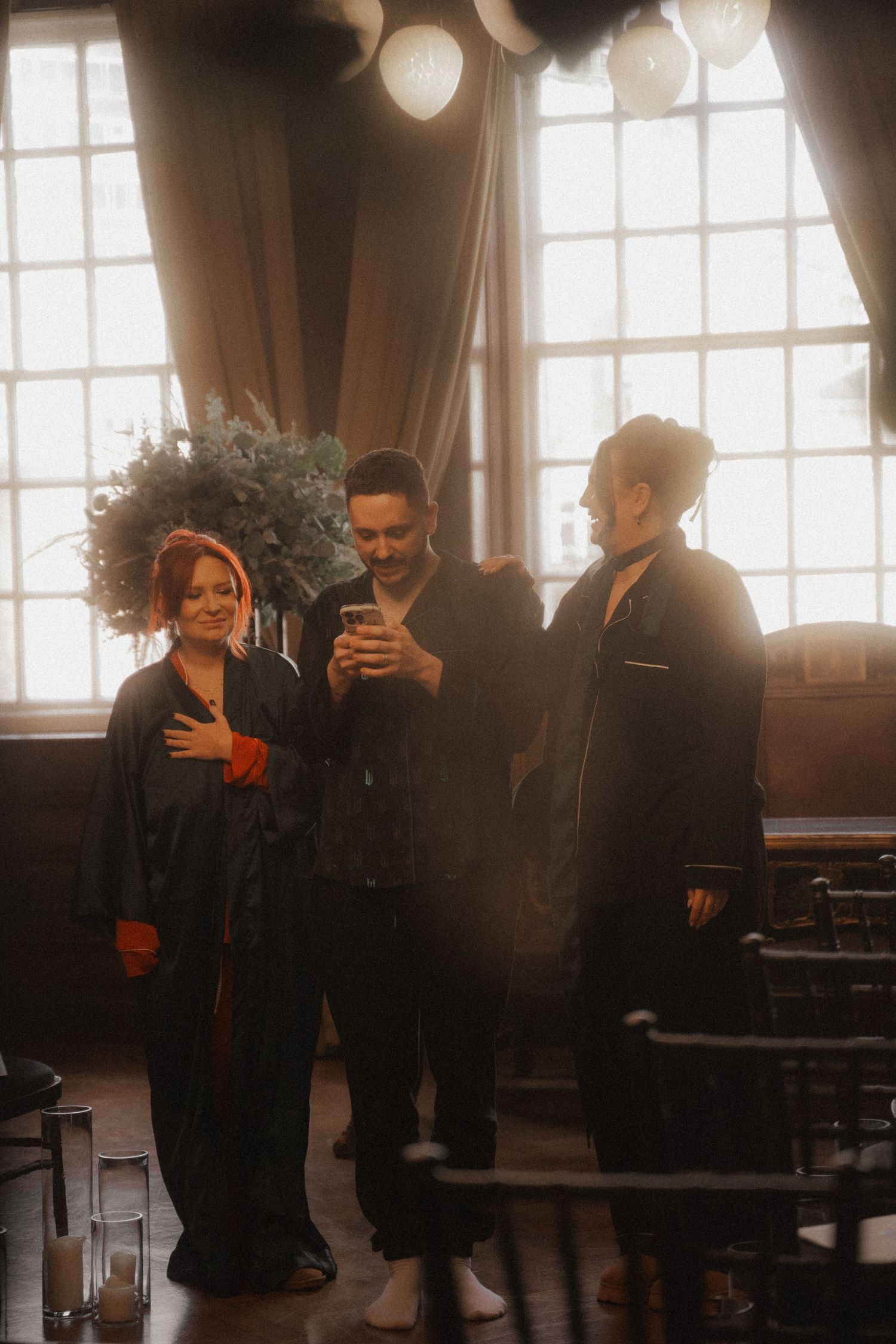 Three people stand together in a warmly lit room with large windows and vintage decor casting a soft sepia-toned ambiance.
