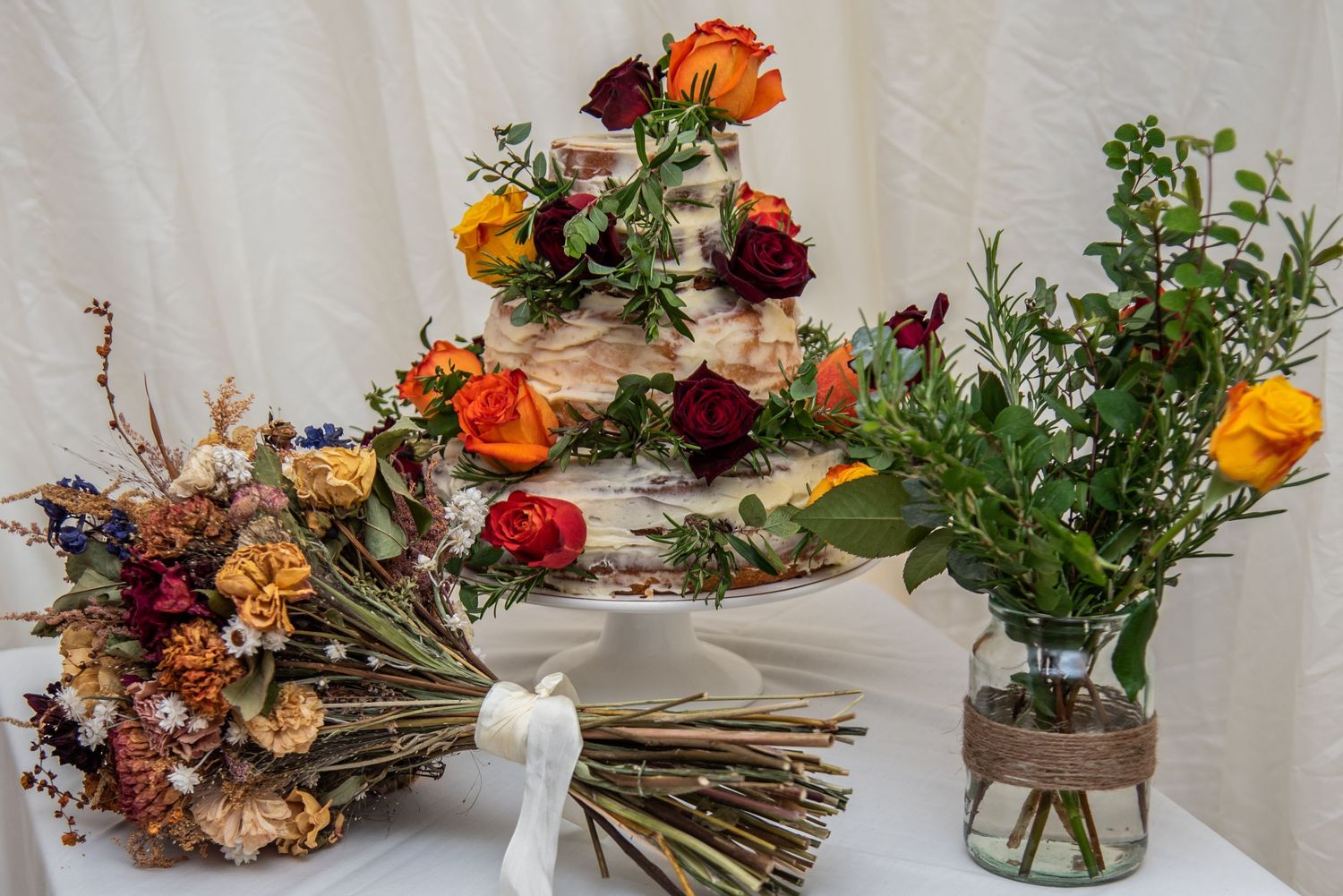 Colorful floral arrangements featuring orange and burgundy roses displayed with dried bouquets against a white wall.