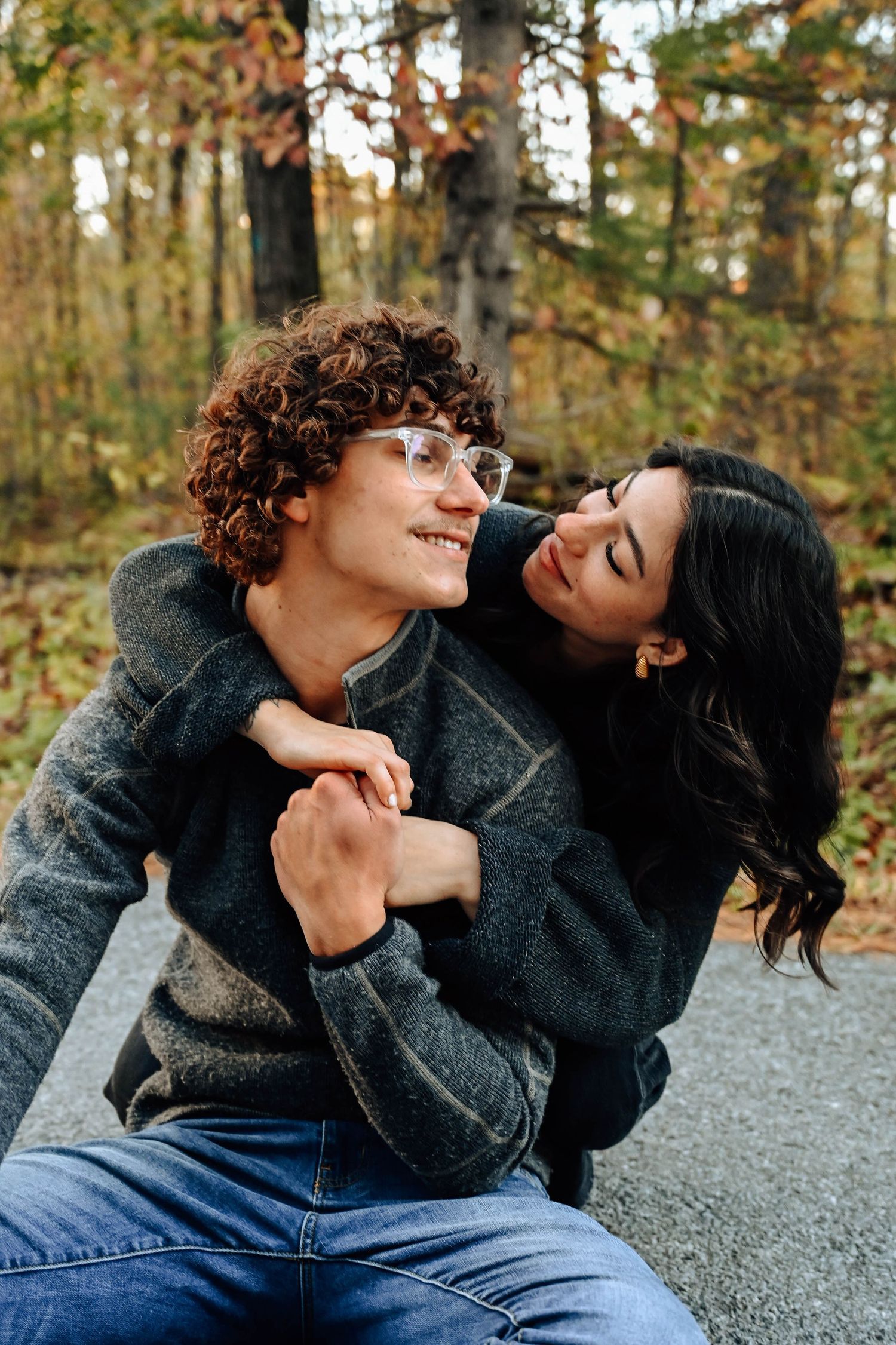 A romantic autumn couple embrace and laugh together outdoors while wearing cozy sweaters in a forest setting.