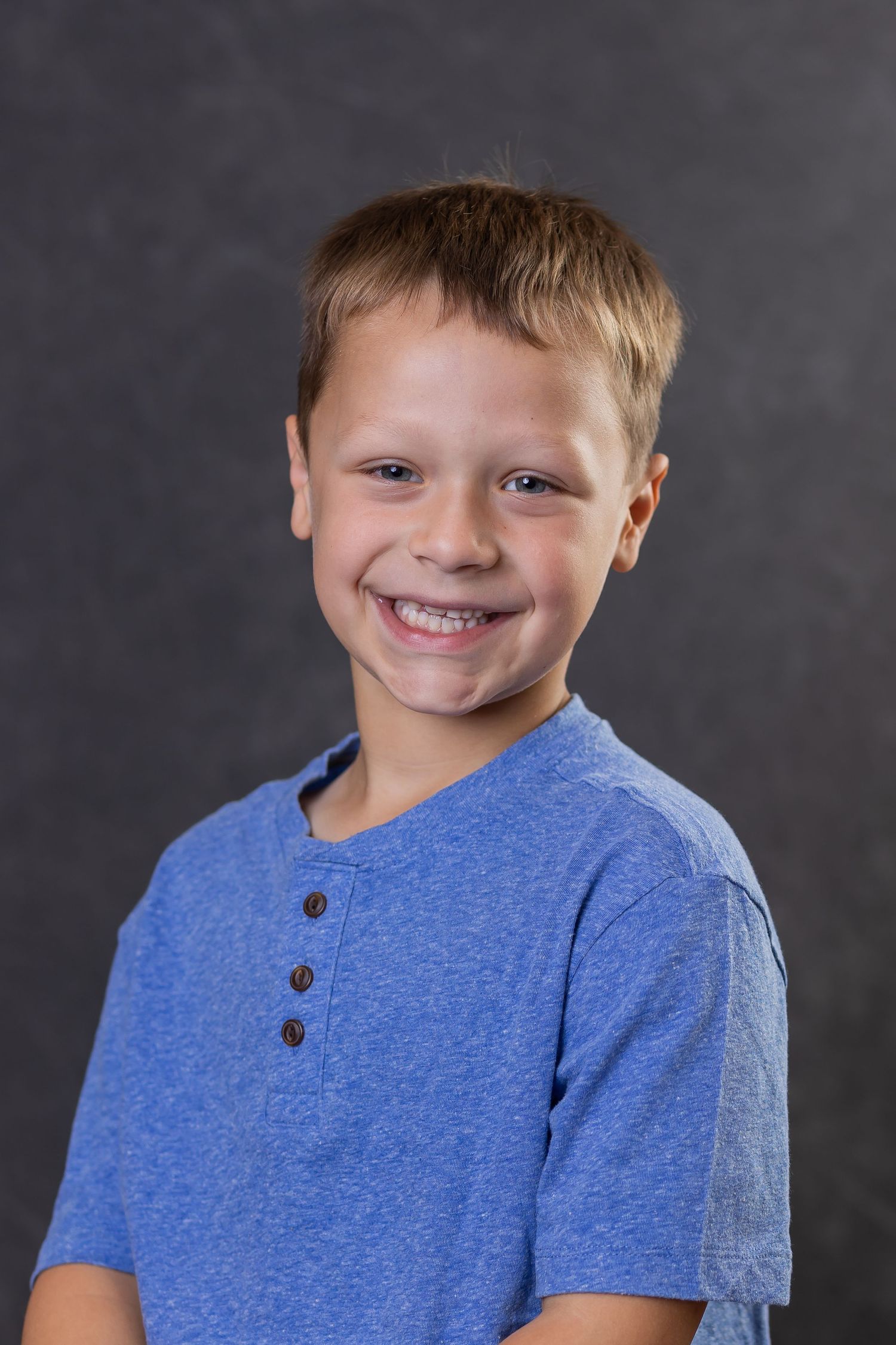 Young student in blue henley shirt smiles warmly against a dark gray background for a school portrait.