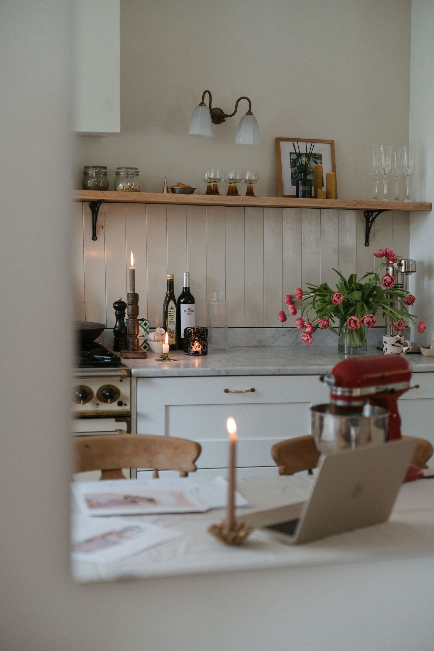 Cozy kitchen scene with candlelit table, laptop, wine bottles, and pink flowers in a rustic white and wood interior.