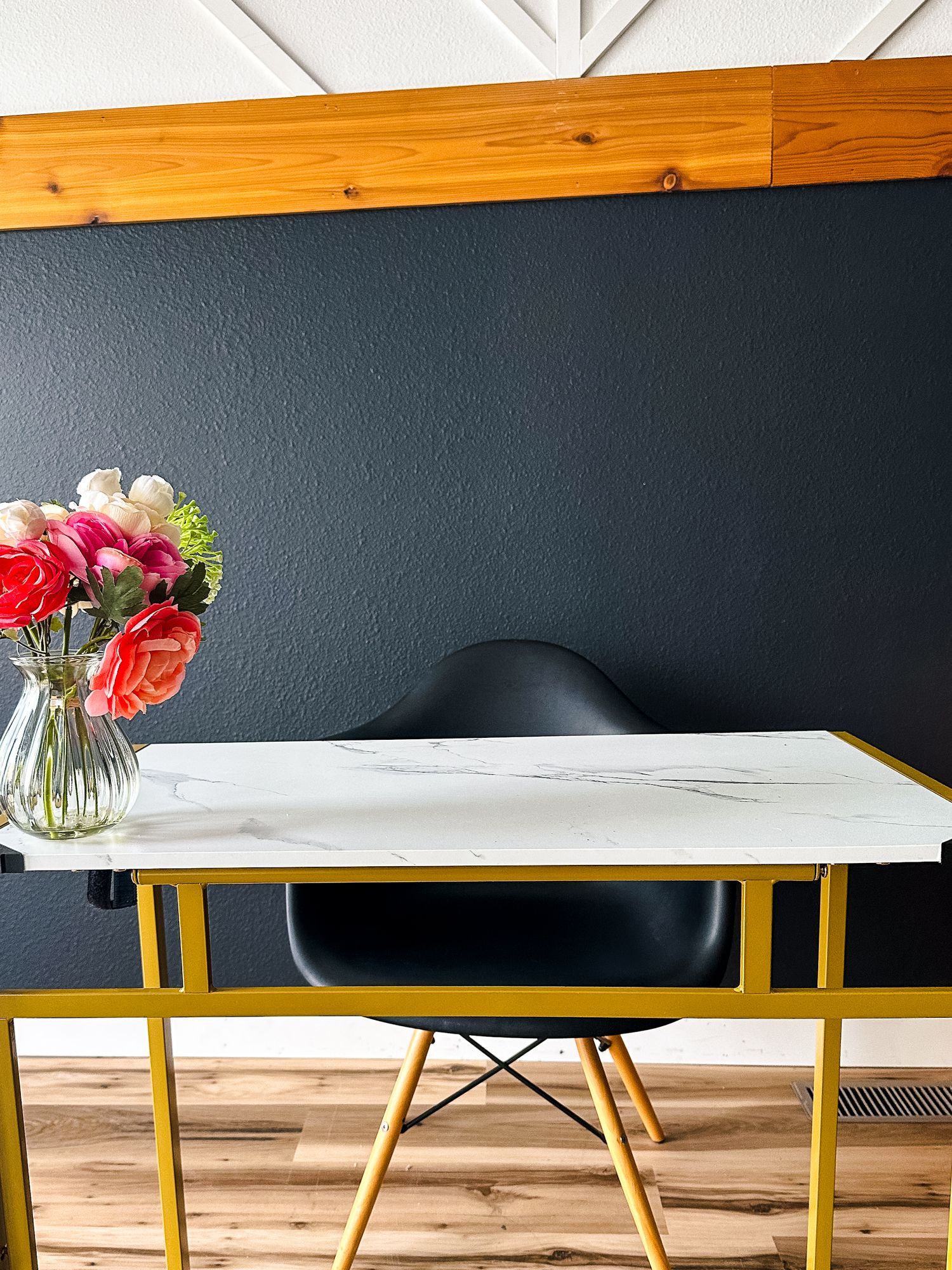 Modern desk setup with marble top table, yellow metal legs, black chair and pink roses in glass vase against navy wall.