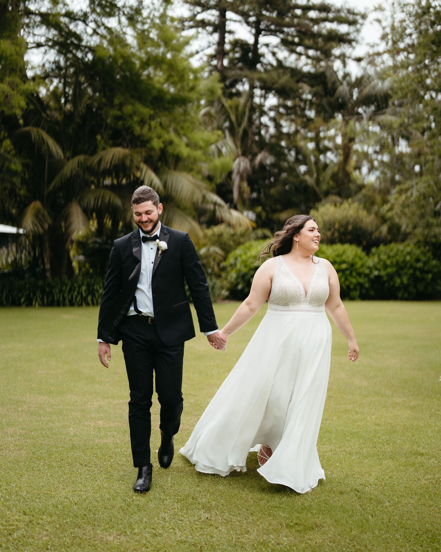 Newlyweds in formal attire walking and holding hands across a lush green lawn surrounded by tropical palm trees.