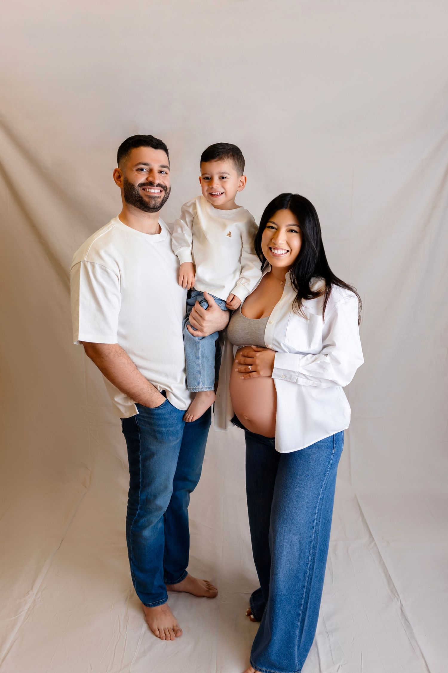 A family of three poses together against a neutral background, with the expecting mother showing her baby bump.