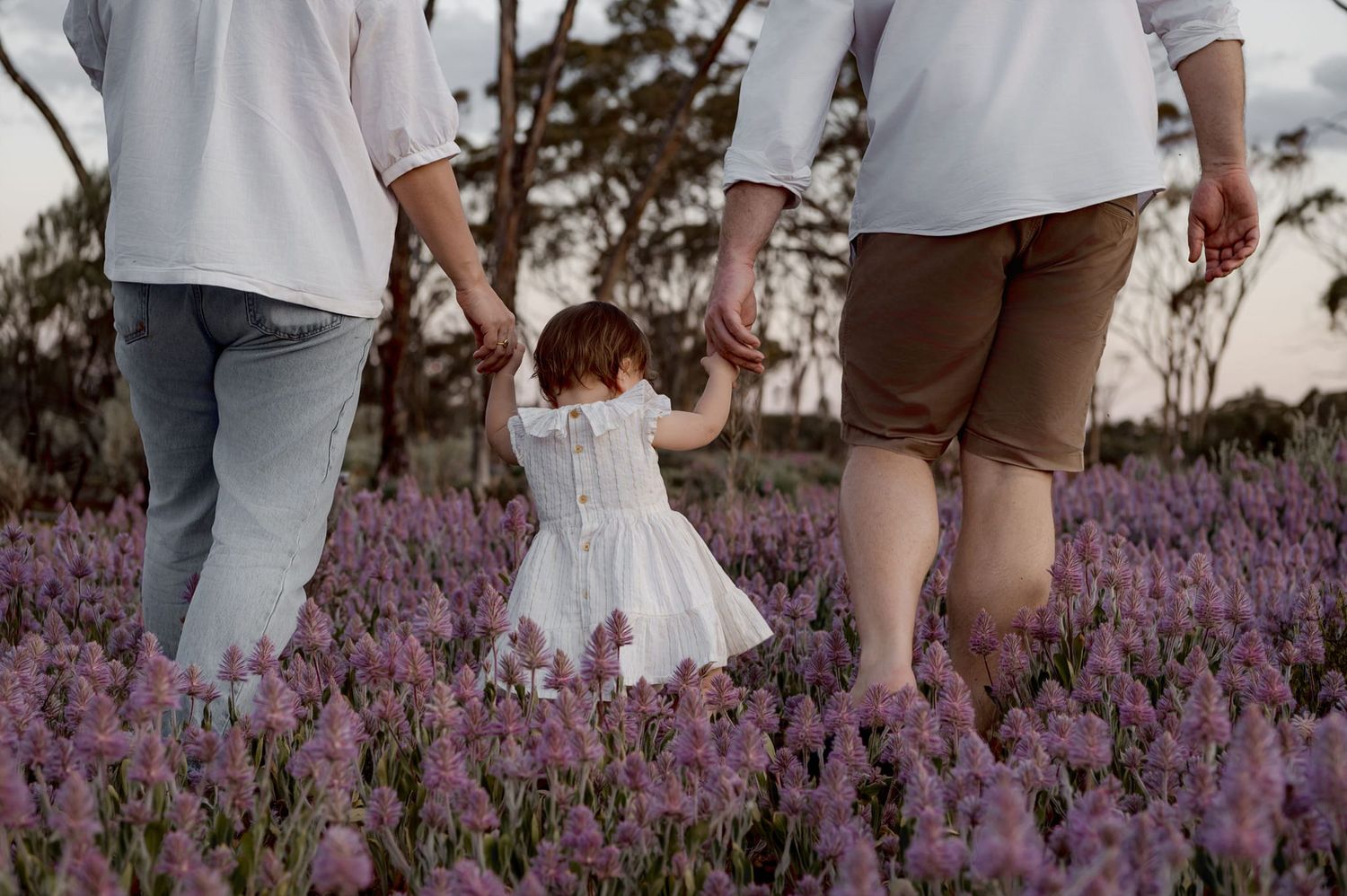 A family stands in a field of purple wildflowers at sunset with their dog while lifting a small child in the air.