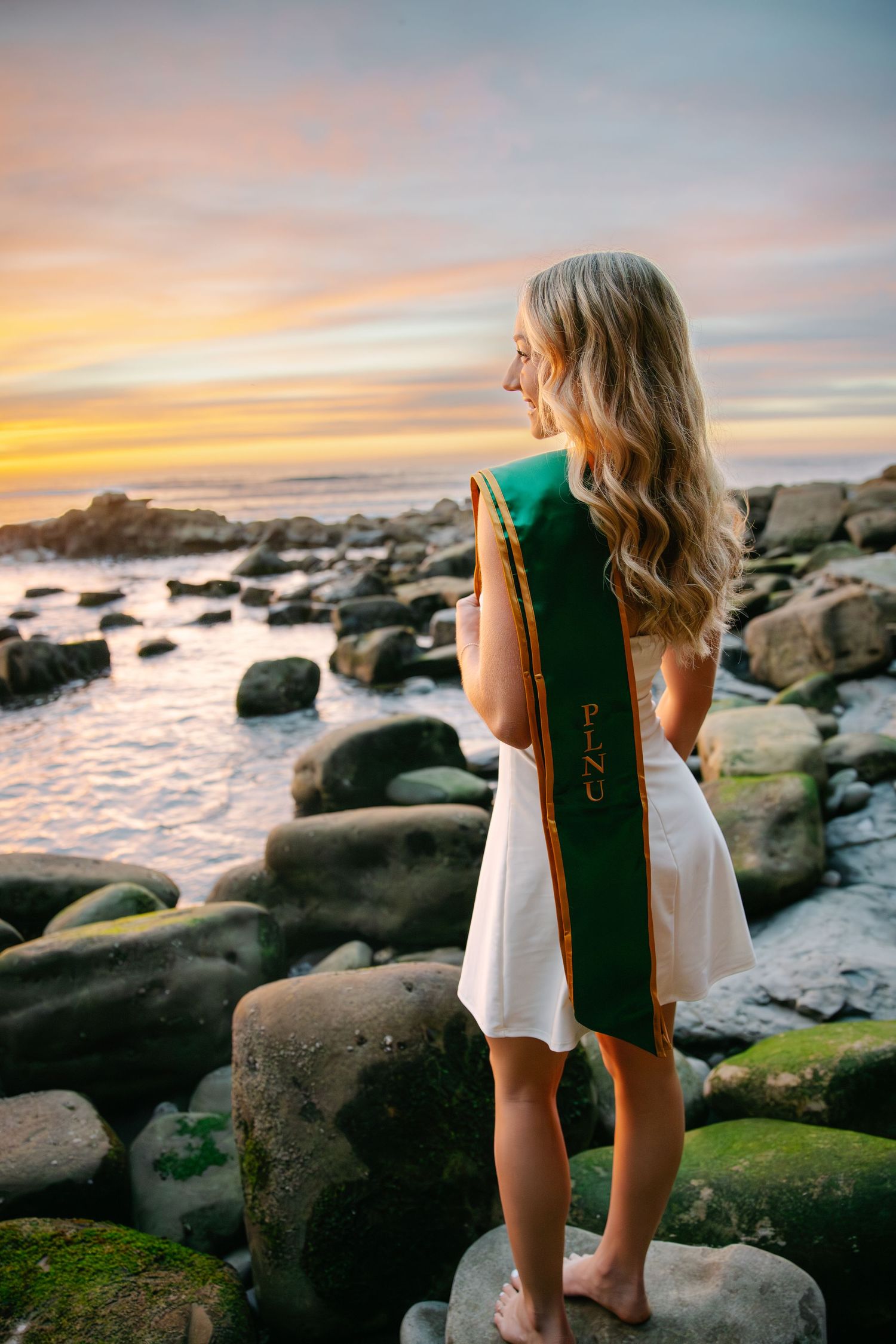 A person in a white dress and green graduation stole stands on rocky coastline at sunset.