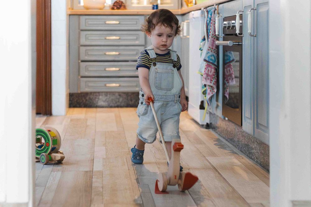 A young child in overalls plays with a wooden toy cleaning set on hardwood floors in a kitchen.