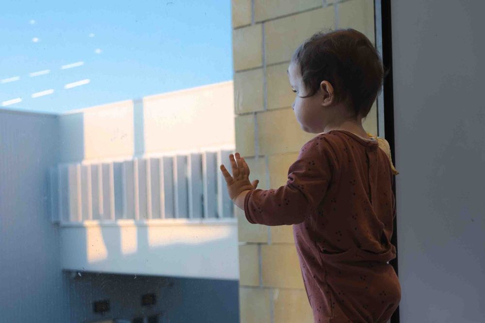 Small child in brown shirt standing near a window looking outside at blue sky and modern architecture.