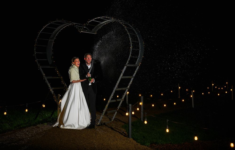 A newlywed couple celebrating with a champagne spray at night, standing under a heart-shaped light arch, captured by SimplyGreatShots Photog