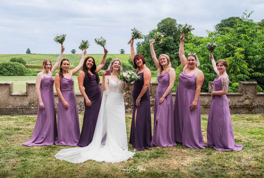 Bride Charlotte and her bridesmaids in elegant purple dresses raising their bouquets in celebration at Fort Henry wedding venue in Rutland,