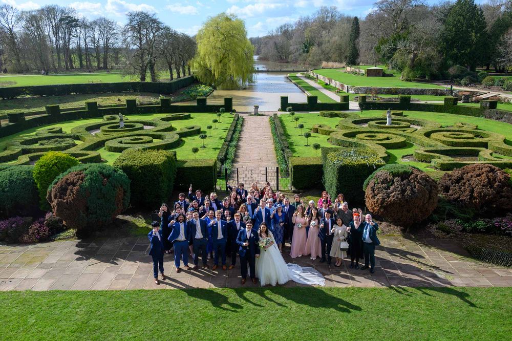 An aerial group photograph of a wedding party raising glasses in a champagne toast, set against the stunning formal parterre gardens and lak