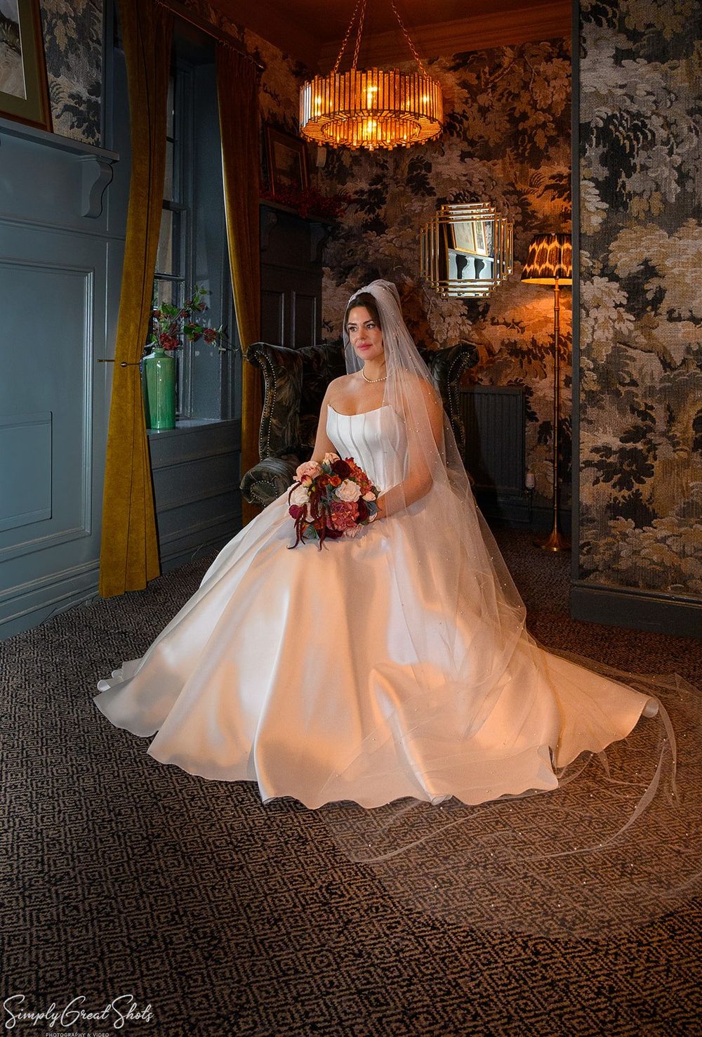 A dramatic full-length portrait of a bride in a sweeping ballgown and long veil, holding an autumnal bouquet, set against the warm ambient l