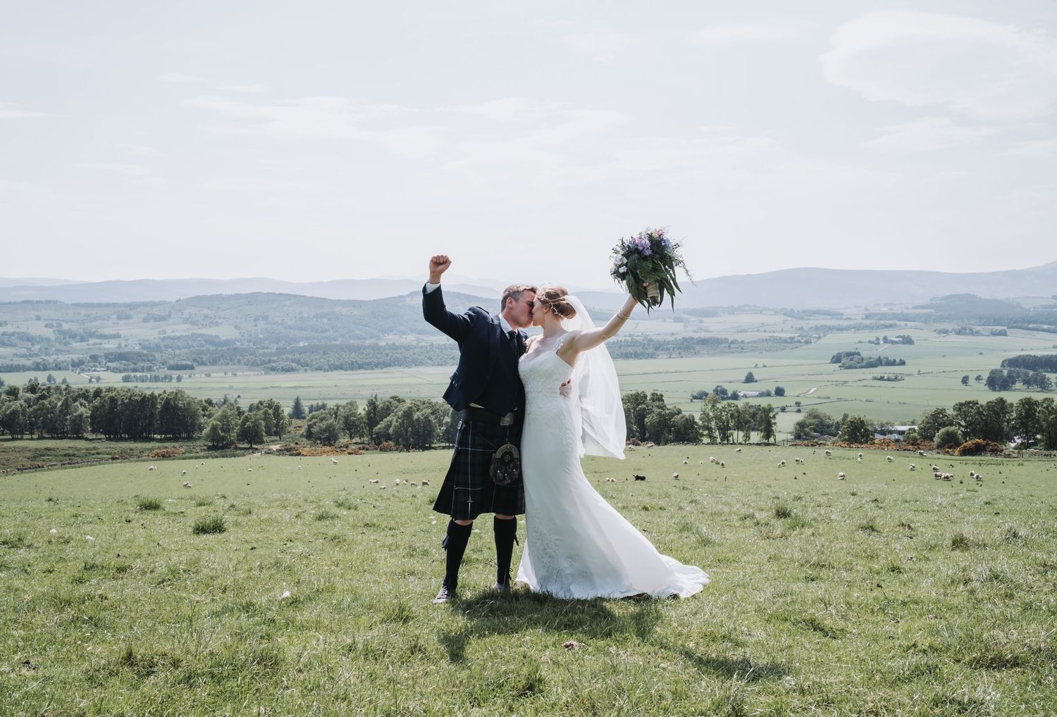 Joyful bride and kilted groom celebrating on a green hilltop with scenic valley views.