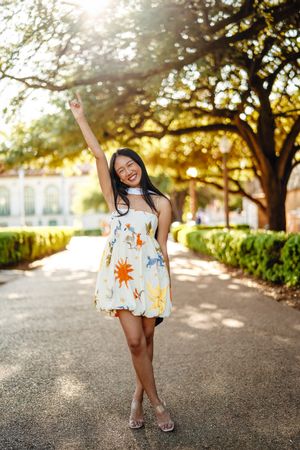 UT Austin grad wearing white sundress with colorful handprint designs twirls on sun-dappled garden path at sunset,