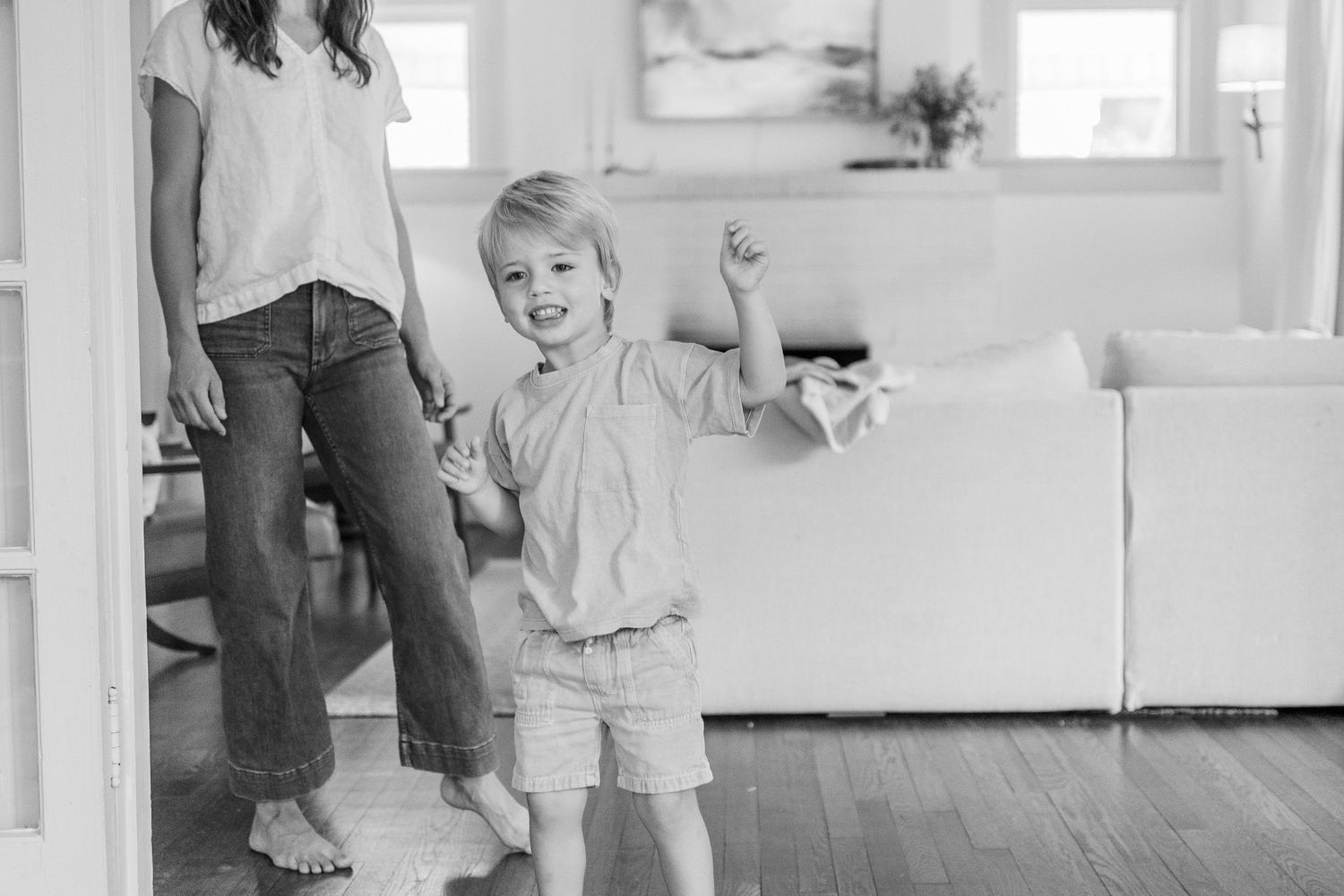 A black and white photo shows an excited young child in shorts and a light shirt standing in a bright living room interior.
