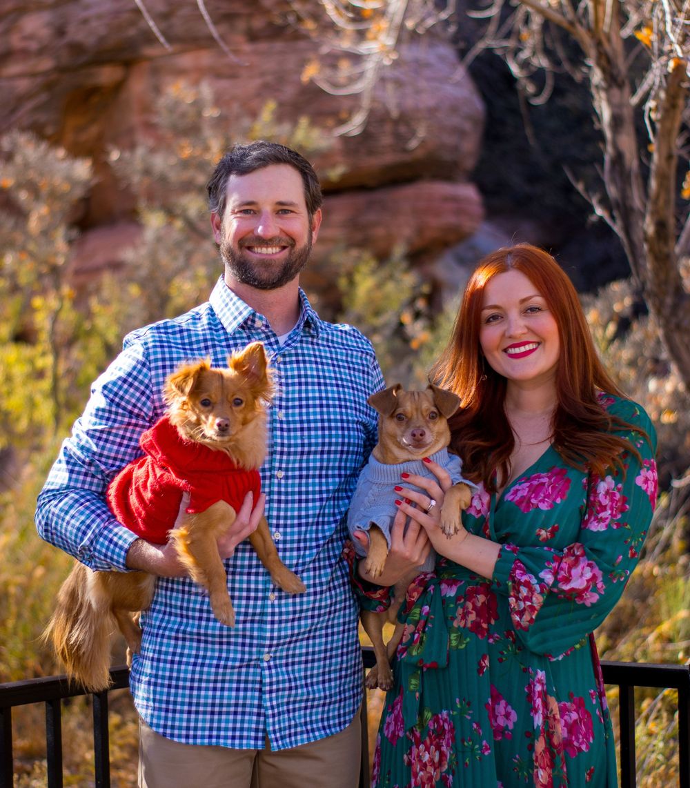 Engaged couple holding two small dogs in colorful sweaters with red rock formation background.