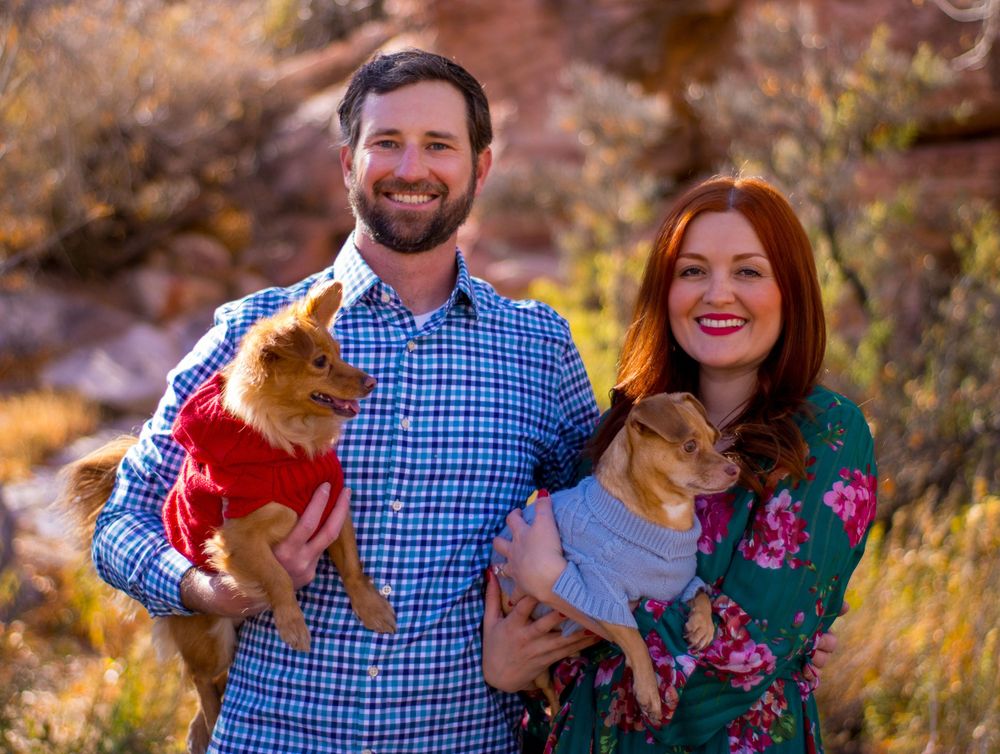 Smiling couple holding two small dogs in red and grey sweaters in desert setting.