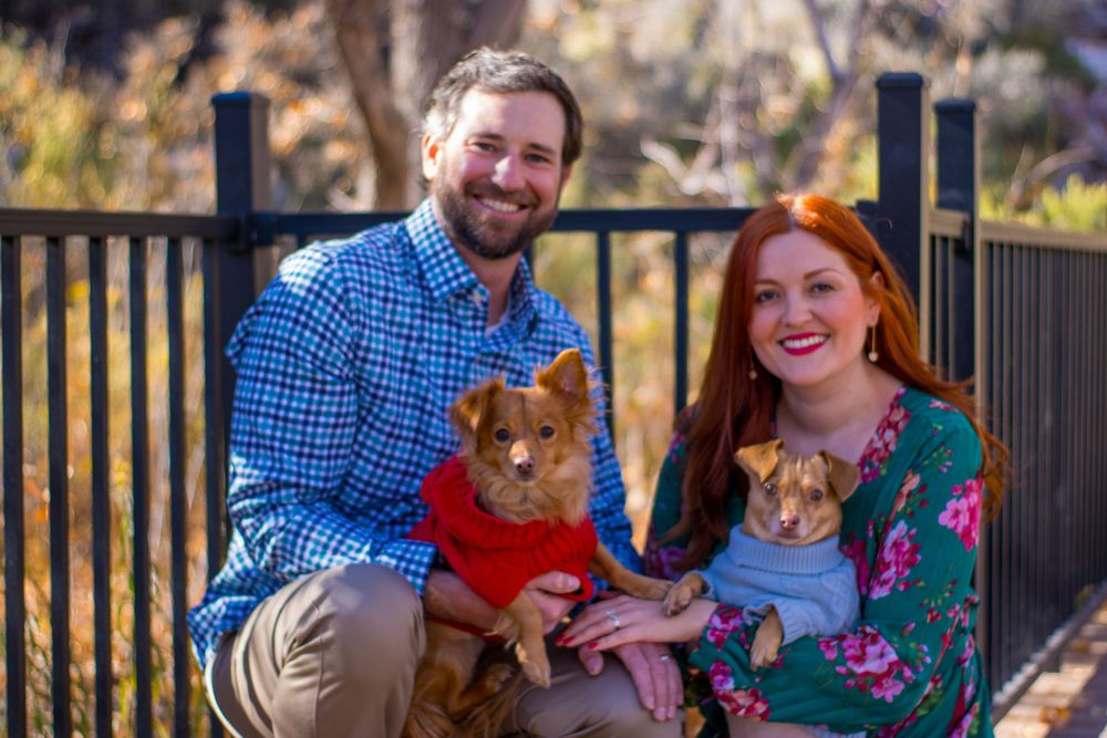 Happy couple kneeling outdoors with two small dogs dressed in red and blue sweaters.