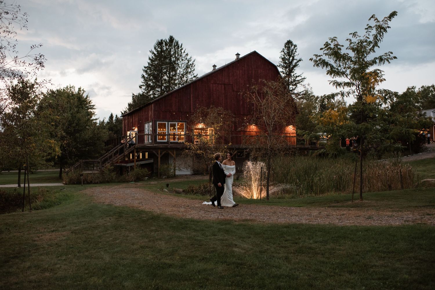 A rustic red barn illuminated at dusk creates a romantic backdrop for a couple embracing on a grassy lawn.