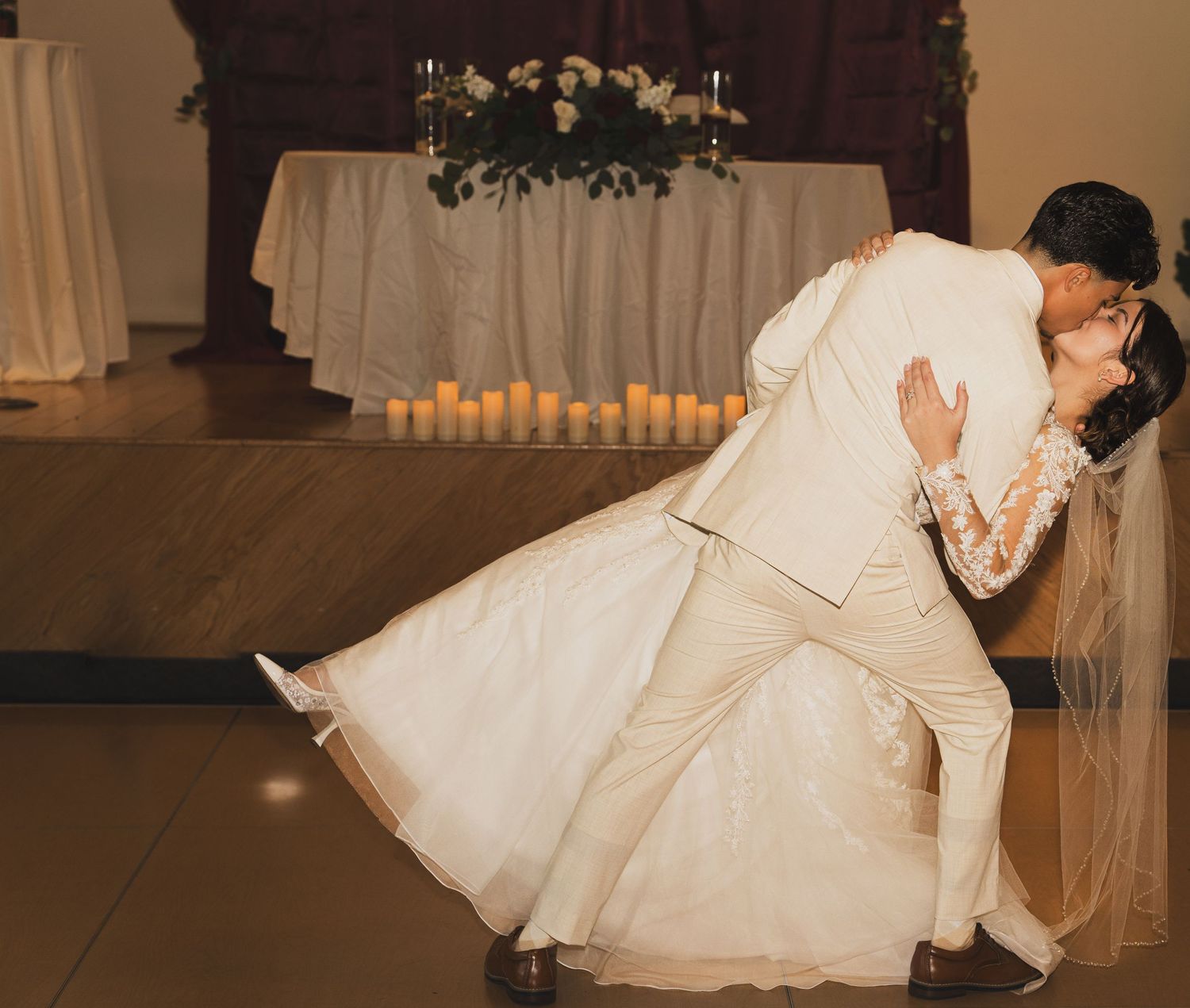 A couple in wedding attire share a romantic dip during their first dance on the reception floor with soft candlelight glowing.