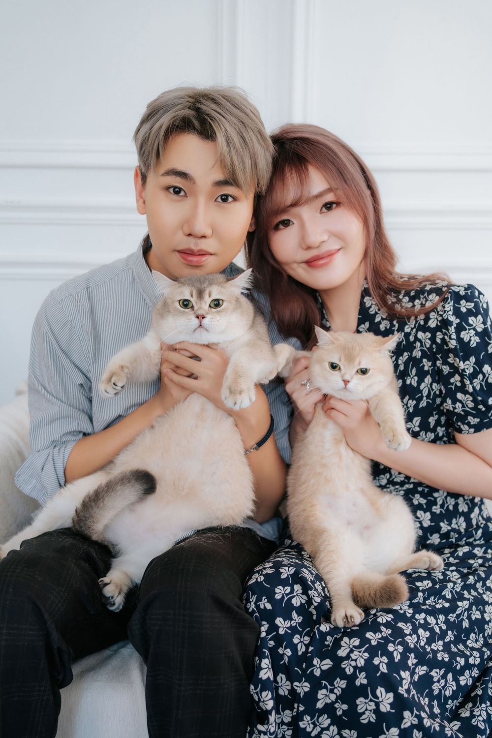Couple posing with their two fluffy white cats against a white wall.