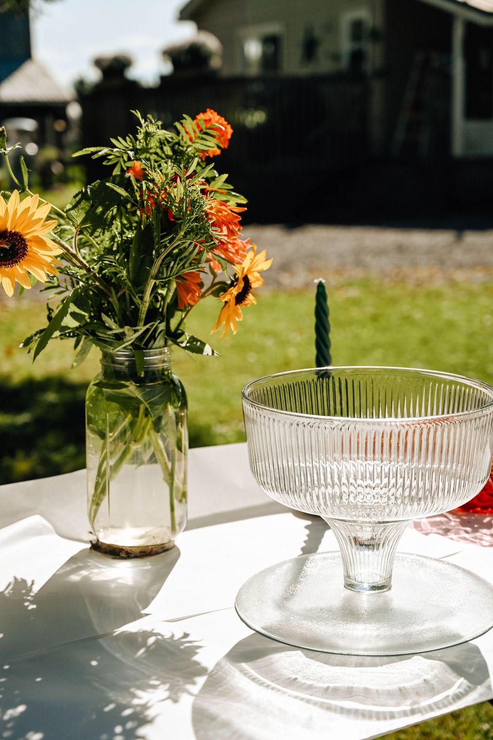 A mason jar with sunflowers and an elegant glass pedestal bowl sit on a white table in bright summer sunlight outdoors.