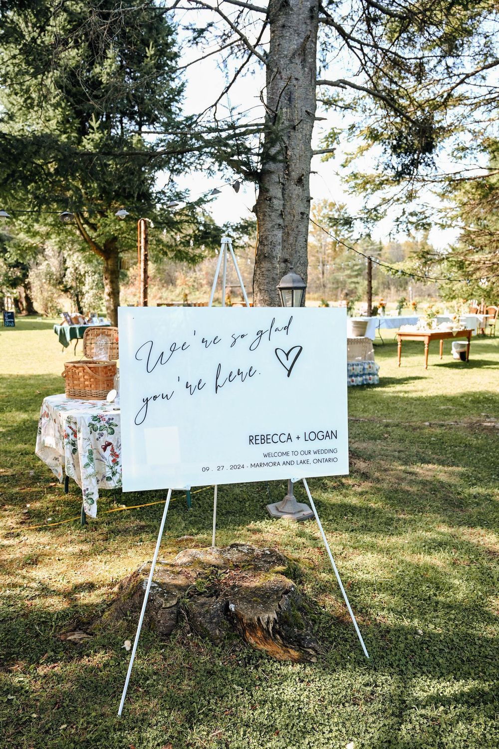 Outdoor wedding welcome sign on an easel with a floral tablecloth and picnic basket in a sunny garden setting.